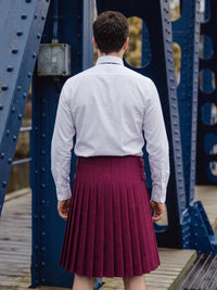A man viewed from behind wearing a white long-sleeve dress shirt and a pleated burgundy kilt, standing on a wooden walkway under a blue steel bridge.