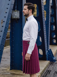 A man wearing a white long-sleeve dress shirt and a burgundy pleated kilt with a black bow tie, standing on a wooden walkway under a blue steel bridge.