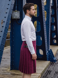 A man in profile wearing a white dress shirt, black bow tie, and a dark burgundy pleated kilt with a black belt, standing on a wooden bridge with a blue steel truss background.