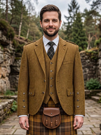 A man wearing a traditional Scottish outfit, featuring a brown corduroy blazer and waistcoat over a white shirt and dark plaid tie, paired with a brown leather sporran and a brown and yellow tartan kilt, set against a blurred outdoor background of stone walls and pine trees.