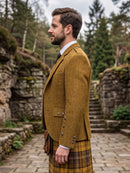 A man in profile wearing a brown tweed blazer, white dress shirt, dark tie, and a yellow-and-brown plaid kilt, standing outdoors on a stone path with trees and stone walls in the background.