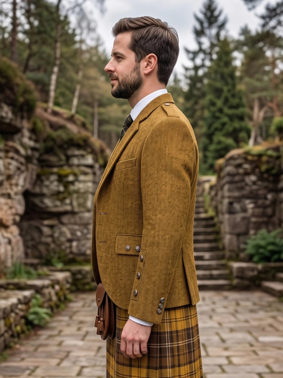 A man in profile wearing a brown tweed blazer, white dress shirt, dark tie, and a yellow-and-brown plaid kilt, standing outdoors on a stone path with trees and stone walls in the background.