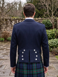 A man viewed from behind wearing a navy blue Scottish formal jacket with silver buttons and a tartan kilt, standing outdoors on a stone path with trees and a stone wall in the background.