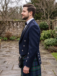 A man in profile wearing a navy blue kilt jacket with silver buttons, a white shirt, a black bow tie, and a green and blue tartan kilt, standing outdoors on a stone path.