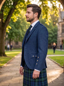 A man wearing a navy blue tweed blazer, white dress shirt, dark tie, and a green and blue plaid kilt stands on a stone pathway in a park with trees and a building in the background.