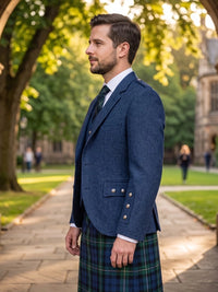 A man wearing a navy blue tweed blazer, white dress shirt, dark tie, and a green and blue plaid kilt stands on a stone pathway in a park with trees and a building in the background.