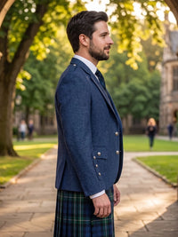 A man in profile wearing a navy blue double-breasted blazer with silver buttons and a green tartan kilt, standing on a stone path in a park with trees and a historic building in the background.