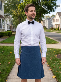 A man wearing a white dress shirt, black bow tie, and a blue pleated kilt stands on a suburban sidewalk.