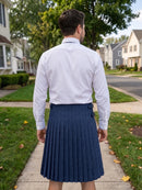 A man viewed from behind wearing a white long-sleeve dress shirt and a navy blue pleated kilt, standing on a suburban sidewalk.