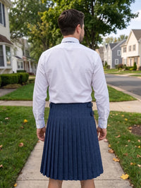 A man viewed from behind wearing a white long-sleeve dress shirt and a navy blue pleated kilt, standing on a suburban sidewalk.