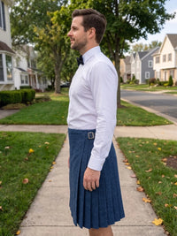 A man standing on a suburban sidewalk wearing a white dress shirt, black bow tie, and a blue pleated kilt with a small silver buckle.