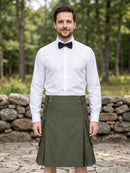 A man wearing an olive green kilt with side pockets, a white dress shirt, and a black bow tie, standing outdoors in front of a stone wall and green forest background.