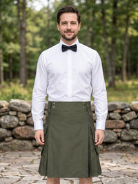 A man wearing an olive green kilt with side pockets, a white dress shirt, and a black bow tie, standing outdoors in front of a stone wall and green forest background.