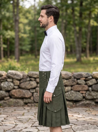 A man wearing a white dress shirt, black bow tie, and an olive green kilt stands in profile outdoors on a stone path with a forest and stone wall in the background.