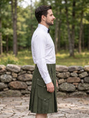 A man wearing a white long-sleeve dress shirt and a dark olive green pleated kilt, standing outdoors in front of a stone wall and a green forest background.