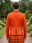 A man viewed from behind wearing a bright orange Scottish kilt jacket with silver buttons and a matching orange and black plaid kilt, standing in a garden setting.