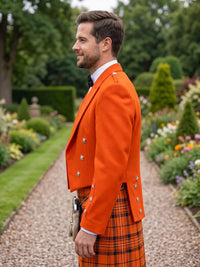 A man in profile wearing a bright orange double-breasted jacket and a matching orange and black plaid kilt, standing on a gravel path in a landscaped garden.