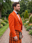 A man standing in a garden wearing a bright orange Scottish jacket and matching orange plaid kilt, with a white shirt, black bow tie, and sporran bag.