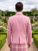 A man viewed from behind wearing a light pink formal jacket with silver buttons and a pink plaid kilt, standing in a garden path.