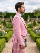 A man in profile wearing a light pink double-breasted blazer, a pink and white plaid kilt, a white shirt, and a black bow tie, standing in a formal garden with trimmed hedges and flower beds.