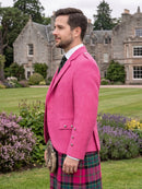 A man in profile wearing a bright pink textured blazer, a red and green tartan kilt, a white shirt, and a black tie, standing outdoors in front of a large stone chateau on a grassy lawn.