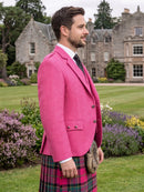 A man in a bright pink herringbone blazer, white dress shirt, black tie, and a red and green tartan kilt with a sporran, standing in a grassy garden with a large stone castle in the background.