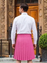 A man viewed from behind wearing a bright pink pleated kilt and a white long-sleeve dress shirt, standing in front of a stone building with a wooden door.