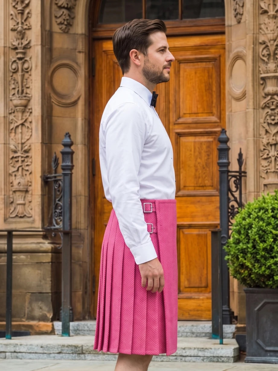 A man in profile wearing a white dress shirt and a bright pink pleated kilt with a matching belt, standing in front of a stone building with a wooden door.