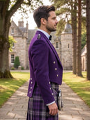 A man in profile wearing a purple Scottish kilt jacket with silver buttons, a purple and black plaid kilt, and a black bow tie, standing outdoors on a stone path with a castle and trees in the background.