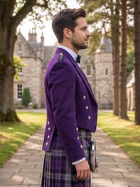 A man in profile wearing a purple Scottish kilt jacket with silver buttons, a purple and black plaid kilt, and a black bow tie, standing outdoors on a stone path with a castle and trees in the background.