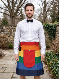 A man wearing a white dress shirt and black bow tie stands outdoors in a garden setting, wearing a rainbow-striped kilt with a matching waist belt and front pocket.