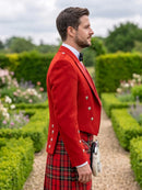 A man in profile wearing a bright red formal jacket and a red tartan kilt, standing in a manicured garden path.