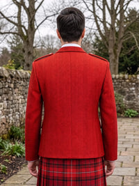 A man viewed from behind wearing a bright red herringbone tweed blazer and a red tartan kilt, standing outdoors on a stone path with trees and a stone wall in the background.