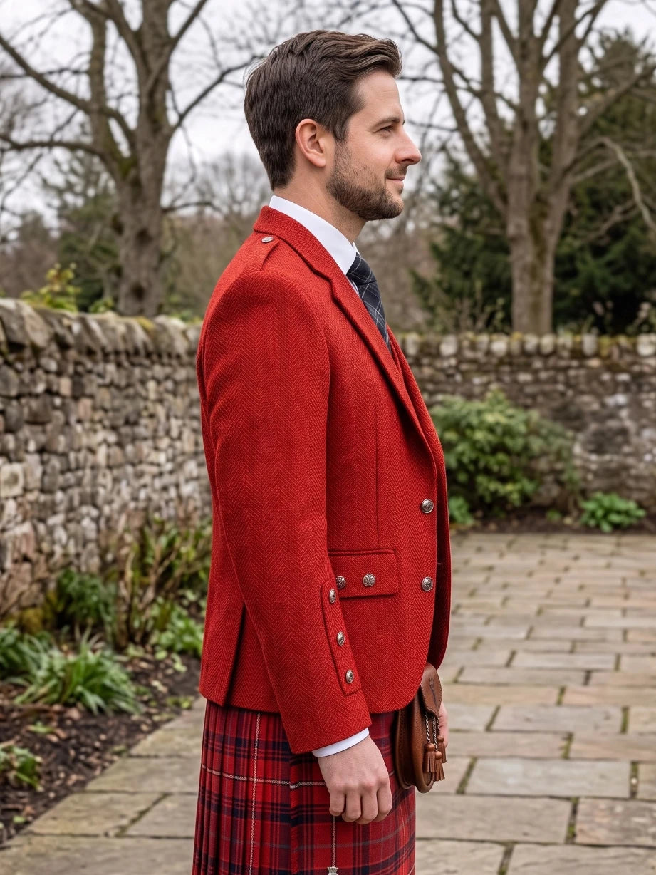 A man in profile wearing a bright red tweed blazer, a red and black tartan kilt, a white shirt, and a dark tie, standing outdoors on a stone path with a stone wall and bare trees in the background.