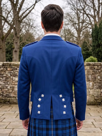 A man viewed from behind wearing a royal blue Scottish kilt jacket with white decorative buttons and a blue and black plaid kilt, standing outdoors in front of a stone wall and bare trees.