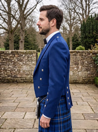 A man in profile wearing a royal blue double-breasted kilt jacket with silver buttons and a blue tartan kilt, standing outdoors in front of a stone wall and bare trees.