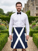 A man wearing a white dress shirt, black bow tie, and a navy blue kilt with a white diagonal cross pattern, standing in a garden path with a stone building in the background.
