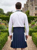 A man viewed from behind wearing a white long-sleeve dress shirt and a navy blue kilt with white wing panels, standing on a garden path with hedges and a stone building in the background.