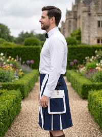 A man standing in profile in a formal garden wearing a white dress shirt, black bow tie, navy blue kilt with white stripes, and a white kilt bag.