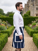 A man in a garden wearing a navy blue and white kilt with a white shirt and black bow tie, standing on a gravel path with hedges and a stone building in the background.