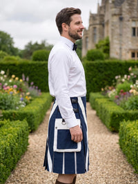 A man in a garden wearing a navy blue and white kilt with a white shirt and black bow tie, standing on a gravel path with hedges and a stone building in the background.