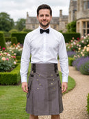 A man wearing a white dress shirt, black bow tie, and a gray kilt with black suspenders and brass buttons, standing in a garden path with a stone building in the background.