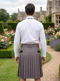 A man viewed from behind wearing a white long-sleeve dress shirt and a pleated gray kilt with side cargo pockets, standing in a landscaped garden path with hedges and flowers in the background.