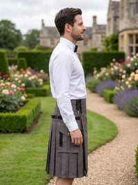 A man in profile wearing a white dress shirt and a dark gray pleated kilt with black straps, standing in a landscaped garden path with hedges and flowers in the background.