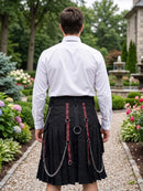A man viewed from behind wearing a black pleated kilt with red and black patterned straps, silver metal rings, and hanging chains, paired with a white dress shirt in a garden setting.