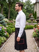 A man in a white dress shirt and black bow tie stands in profile outdoors wearing a black kilt with a red patterned pocket and chain detail, set against a garden background.