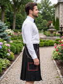 A man in a garden wearing a black kilt with red trim and a white dress shirt, shown in profile. The background features a gravel path, green hedges, and a stone building.