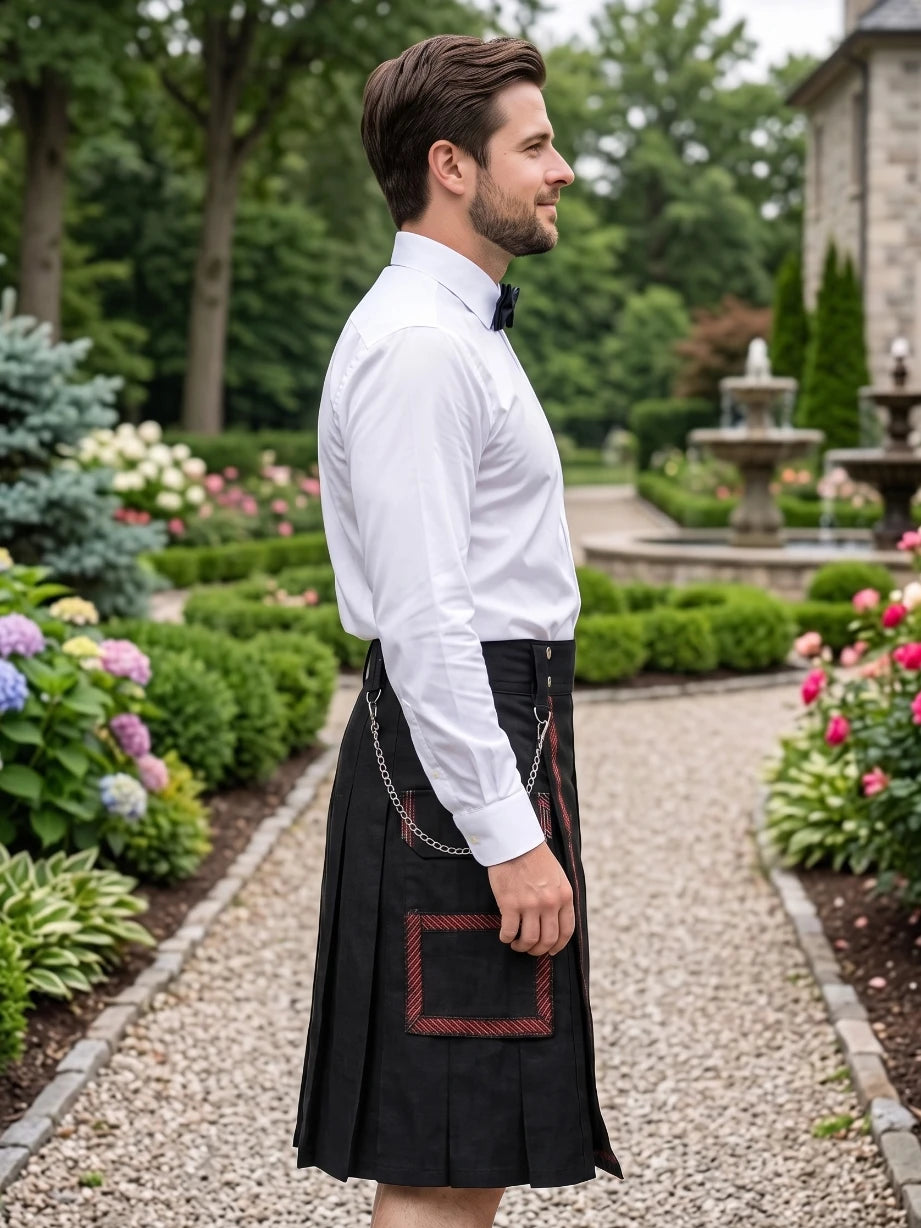 A man in a garden wearing a black kilt with red trim and a white dress shirt, shown in profile. The background features a gravel path, green hedges, and a stone building.