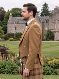 A man in profile wearing a traditional Scottish kilt outfit with a tan jacket, white shirt, black bow tie, and brown plaid kilt, standing in a garden with a large stone mansion in the background.
