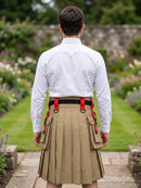 A man viewed from behind wearing a white long-sleeve shirt, a beige pleated kilt with red suspenders and side pockets, and a black belt, standing in a garden pathway.